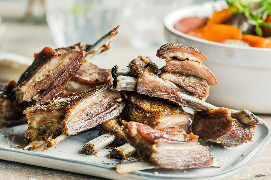 Stekt lammeribbe delt opp og servert på lyst avlangt fat. Hvit skål med glaserte grønnsaker i bakgrunnen.