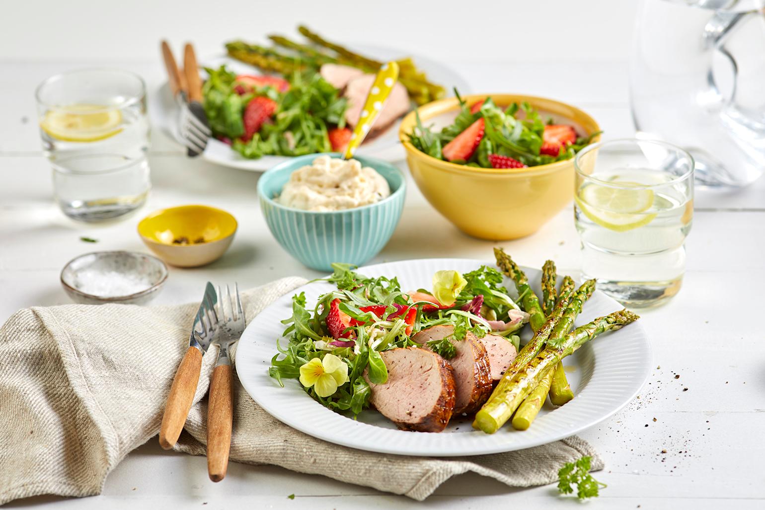 Indrefilet av svin med grillet asparges og sommersalat med jordbær servert på hvit tallerken på dekket hvitt trebord. Små skåler aioli, sommersalat, salt og pepper i bakgrunnen.