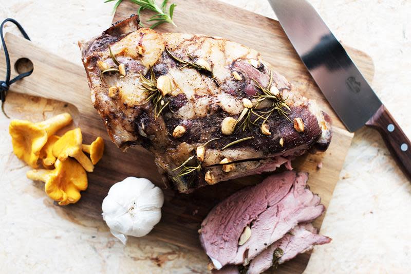 Rosastekt lammelår spekket med hvitløksfedd og fersk rosmarin, servert på trefjøl. Hvitløk, kantareller og fersk rosmarin rundt.