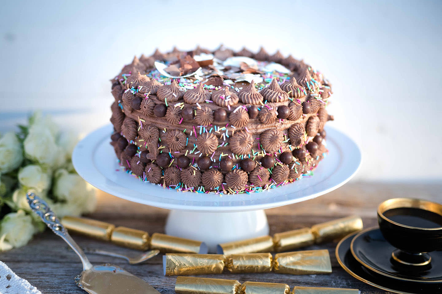 Sjokoladekake til fest med tuttifruttistrø og sjokoladekrem, servert på hvitt stettfat. Kaffekopp, hvite roser og smellbongbonger rundt.