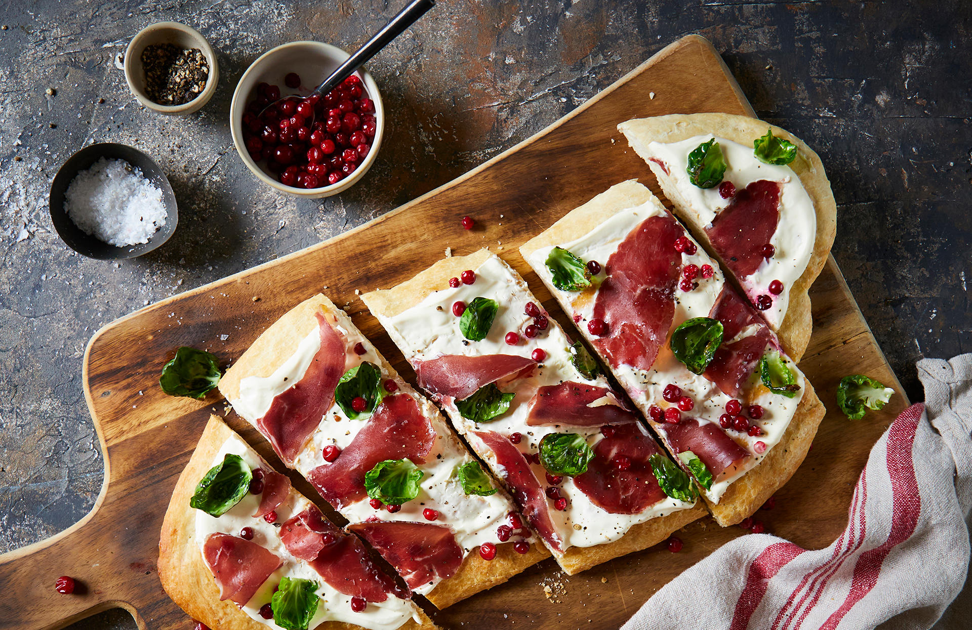 Hvit pizza med fenalår og tyttebær, skåret i skiver og toppet med rosenkålblader. Pizzaen er avlang, og servert på en trefjøl, på rustikk steinbakgrunn. Små skåler med salt, pepper og tyttebær ved siden av.