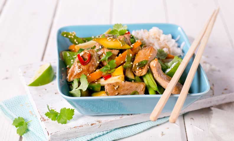 Wok med svinefilet og chili, og fargerike grønnsaker. Servert i blå firkantet bolle med ris og et dryss koriander, på hvitt trebord.