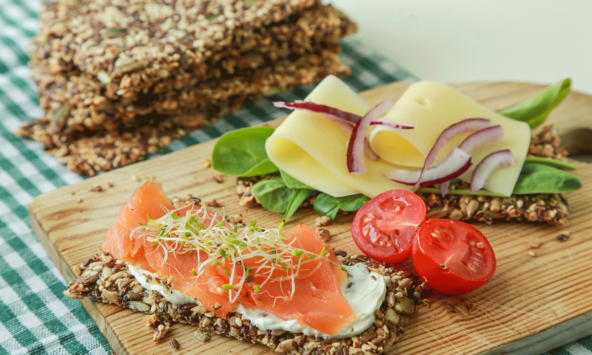 Sprø knekkebrød med kullsyre servert på trefjøl med godt pålegg. Ett knekkebrød med kremost, røkelaks og spirer, og ett med spinat, gulost og rødløk.