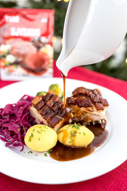 Biter av ribbe på hvit tallerken, med poteter, rosenkål og rødkål. Sjysaus helles over tallerkenen fra et hvitt sausenebb. Pose med Toro sjysaus i bakgrunnen. Rød duk.