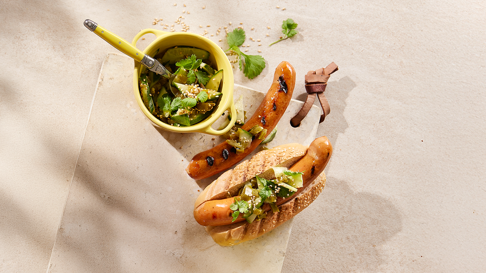 Spicy agurksalat med jalapeño servert over pølse i brød på en beige marmorfjøl med en pølse uten brød ved siden av. Liten gul skål med agurksalat på siden.