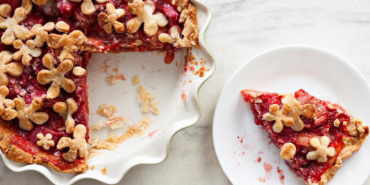 Rabarbrapai i hvit paiform på hvitt marmorbord. Paien er pyntet med små blomster av paideig, og ved siden av står en asjett med et stykke pai.