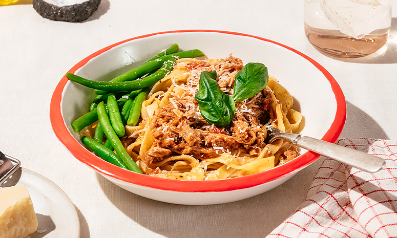 Pasta med pulled pork og aspargesbønner, toppet med basilikum og revet parmesan servert i en dyp hvit skål med en gaffel