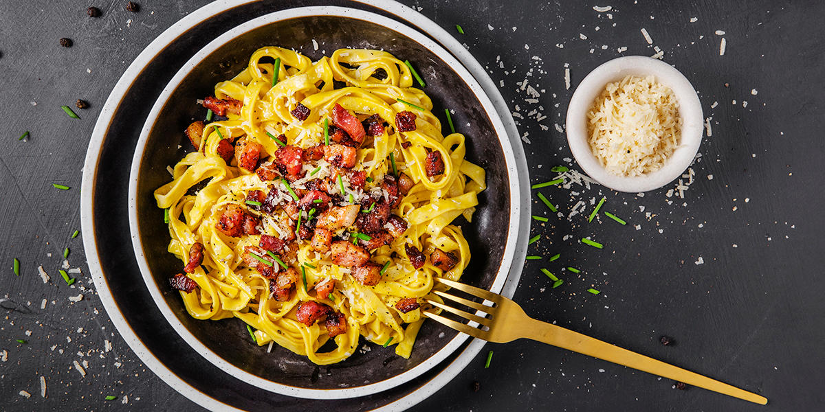 En rykende fersk tallerken med pasta carbonara på mørk bakgrunn, toppet med sprøstekt bacon og revet parmesan.