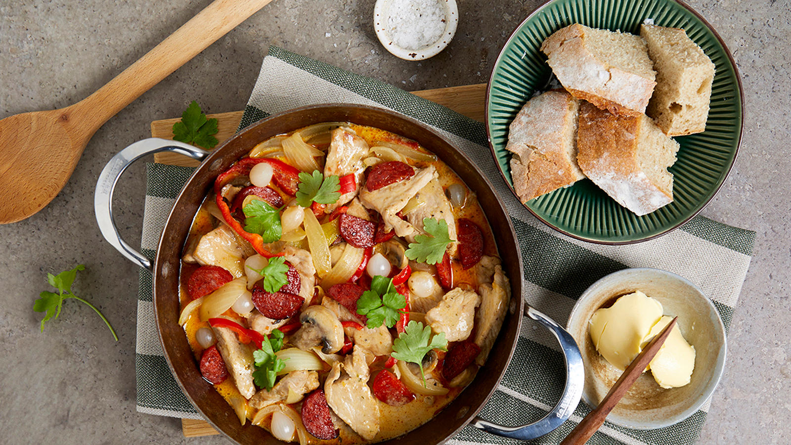 Rask kremet gryte med svinekjøtt og chorizo servert på grønnstripete kjøkkenhåndkle på trefjøl. Grønn skål med brød, og liten lys skål med smør ved siden av.