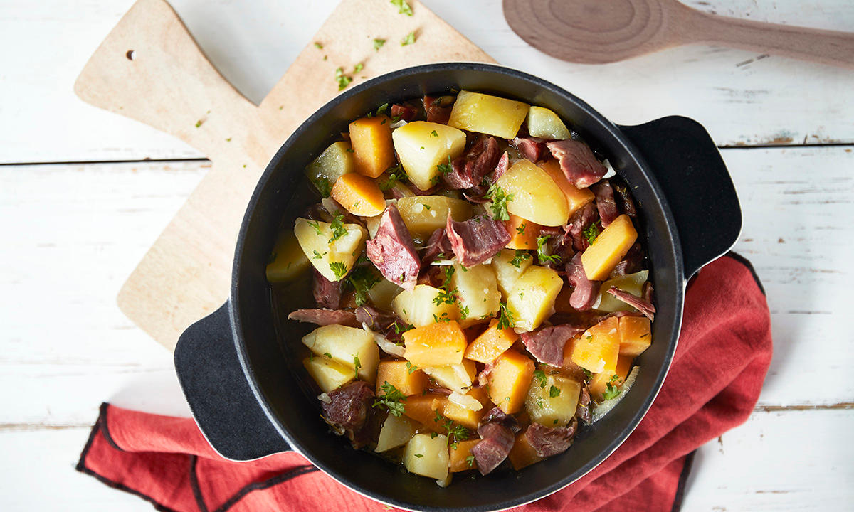 Lapskaus av pinnekjøttrester med potet og kålrabi servert i svart gryte på lys trefjøl med rød linserviett. Tresleiv ved siden av.