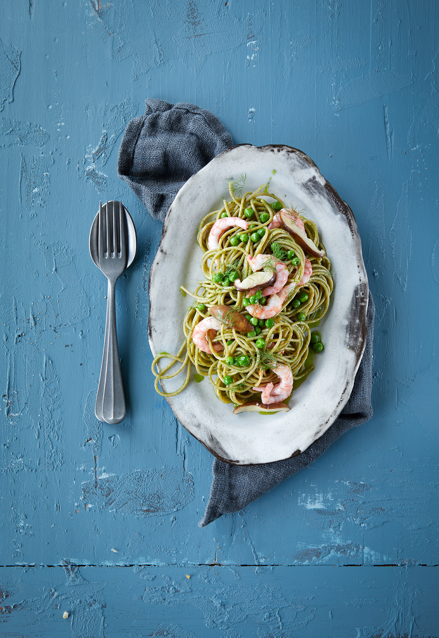 Grønn spaghetti med reker. Spaghetti med reker, erter, sopp og vårløk, servert på oval tallerken på blå bakgrunn.