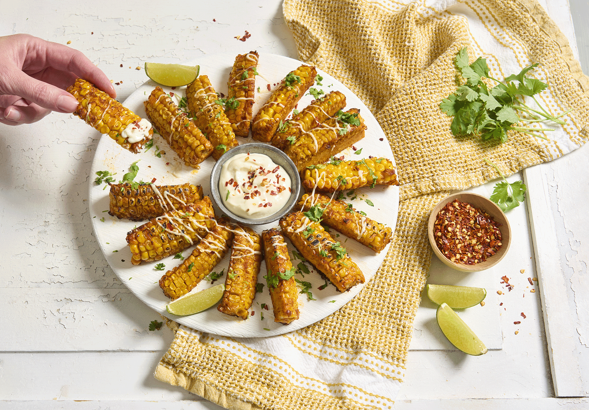 Corn ribs - sommerens diggeste mais, servert på rundt hvitt fat med hakket koriander og limebåter, og en skål dip i midten. En hånd holder en maisrib med litt dip på. Skål med chiliflak på siden.