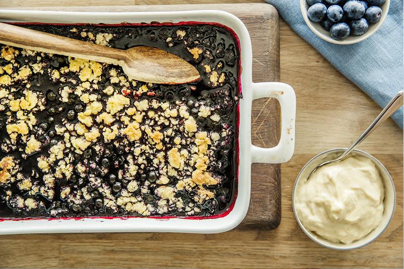 Smuldrepai med blåbær, laget i hvit ildfast form, servert på trefjøl med vaniljekrem. Blå linserviett og ferske blåbær ved siden av.