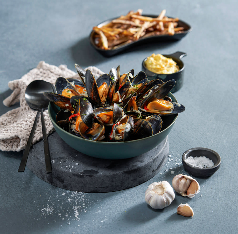 Moules frites - dampede blåskjell og pommes frites servert i skåler, med hjemmelaget aioli i liten skål ved siden av. Hel hvitløk og liten skål med salt i forgrunn.