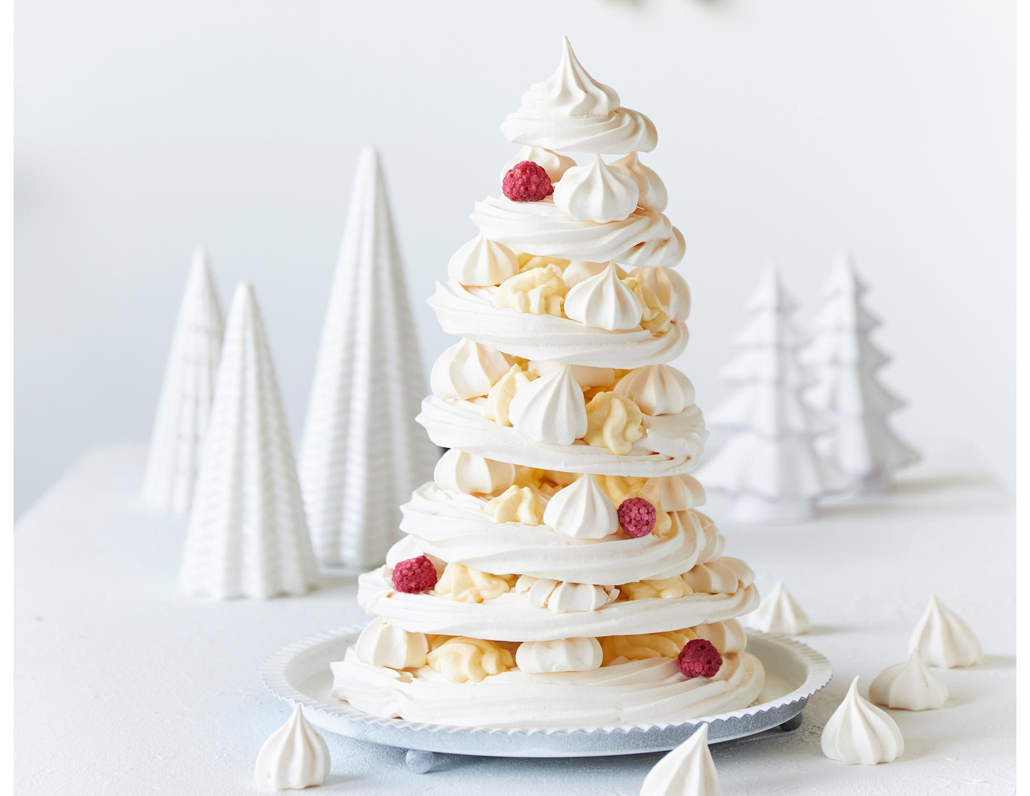 Marengsjuletre med hjemmelaget vaniljekrem; runde skiver av marengs, stablet i form av et juletre med vaniljekrem, friske bringebær og små marengs i mellom lagene. Marengsjuletreet står på hvitt bord med hvite dekortrær i bakgrunnen.