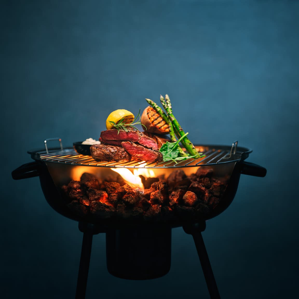 Skyggen av en Grill Perfekt grill med biff, søtpotet, asparges og sitron på flammene. Blåsvart bakgrunn.