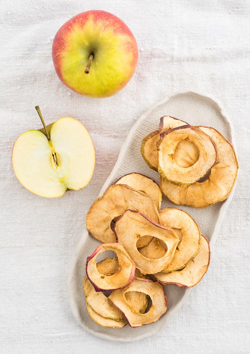 Eplechips med kanel servert på hvitt avlangt fat, på hvit duk. frist rødt og gult eple ved siden av.