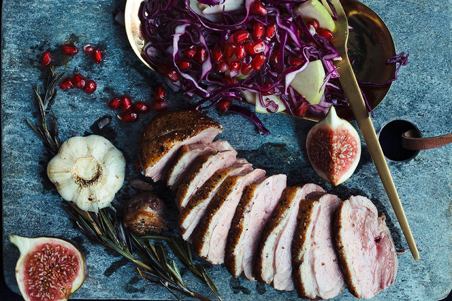 Andebryst servert i skiver med halve fiken, ferske urter, granateplefrø og fruktig rødkålsalat med grønt eple. Gullfarget bestikk ved siden av.