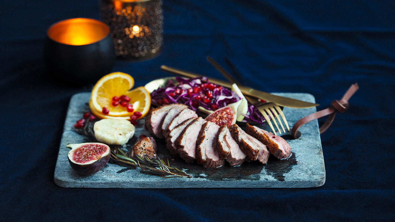 Andebryst med fruktig rødkålsalat, appelsin og fiken servert i skiver på grå steinfjøl. Tente lys i bakgrunnen.