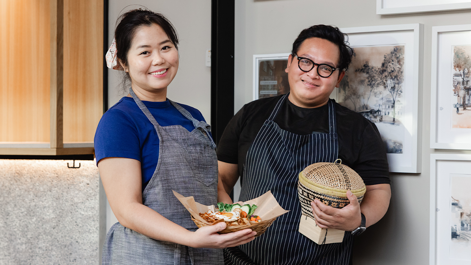 Portrett av to smilende personer, en kvinne og en mann, som begge har på seg forklær over mørke t-skjorter, stående i en restaurant. Kvinnen holder et flettet fat med en indonesisk risrett. Mannen holder en flettet kurv (en boboko) og ser mot kameraet.
