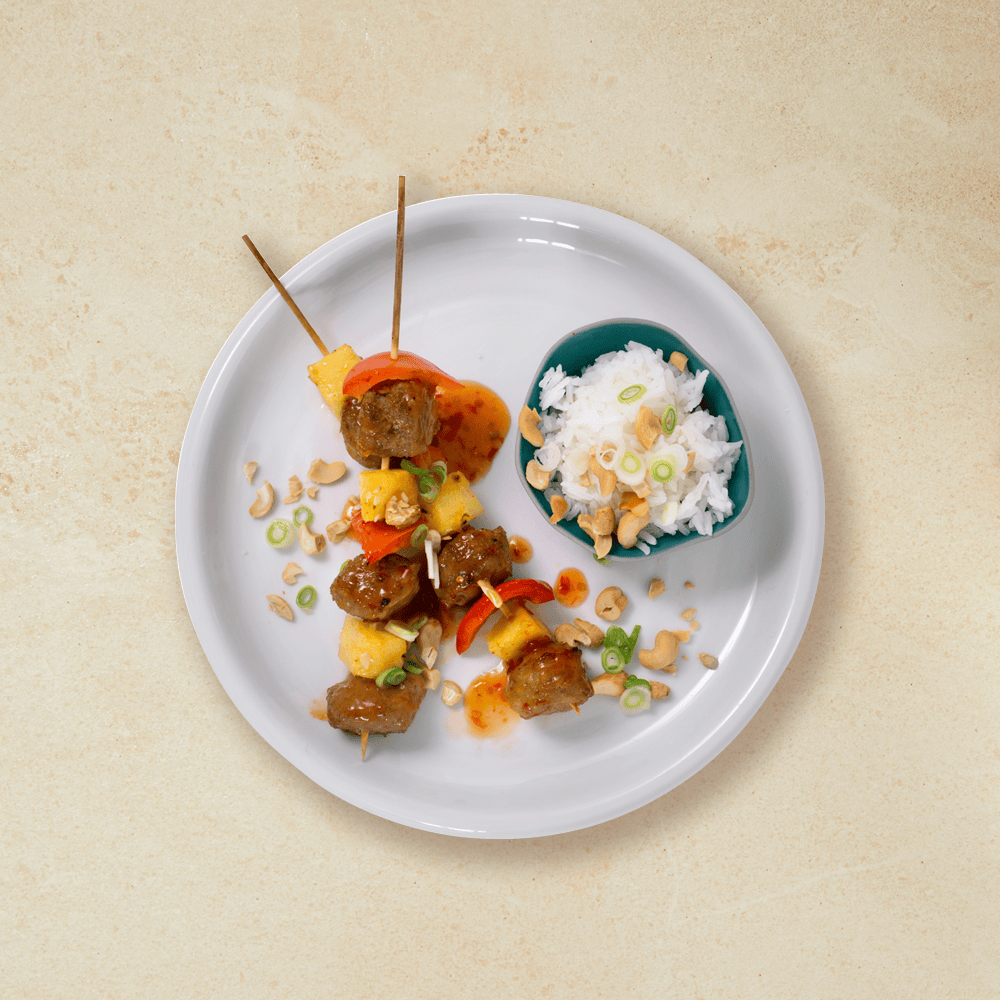 Kyllingkjøttboller på spyd med ananas og paprika, servert med ris i liten blå skål, på grå tallerken.