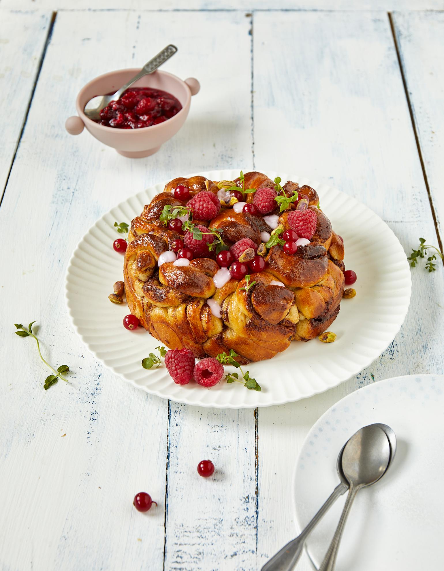 Kanelbollekake med rips og bringebær på toppen, pyntet med friske urter, og servert på hvitt fat på hvitt trebord, med en rosa skål rørte bringebær i bakgrunnen.