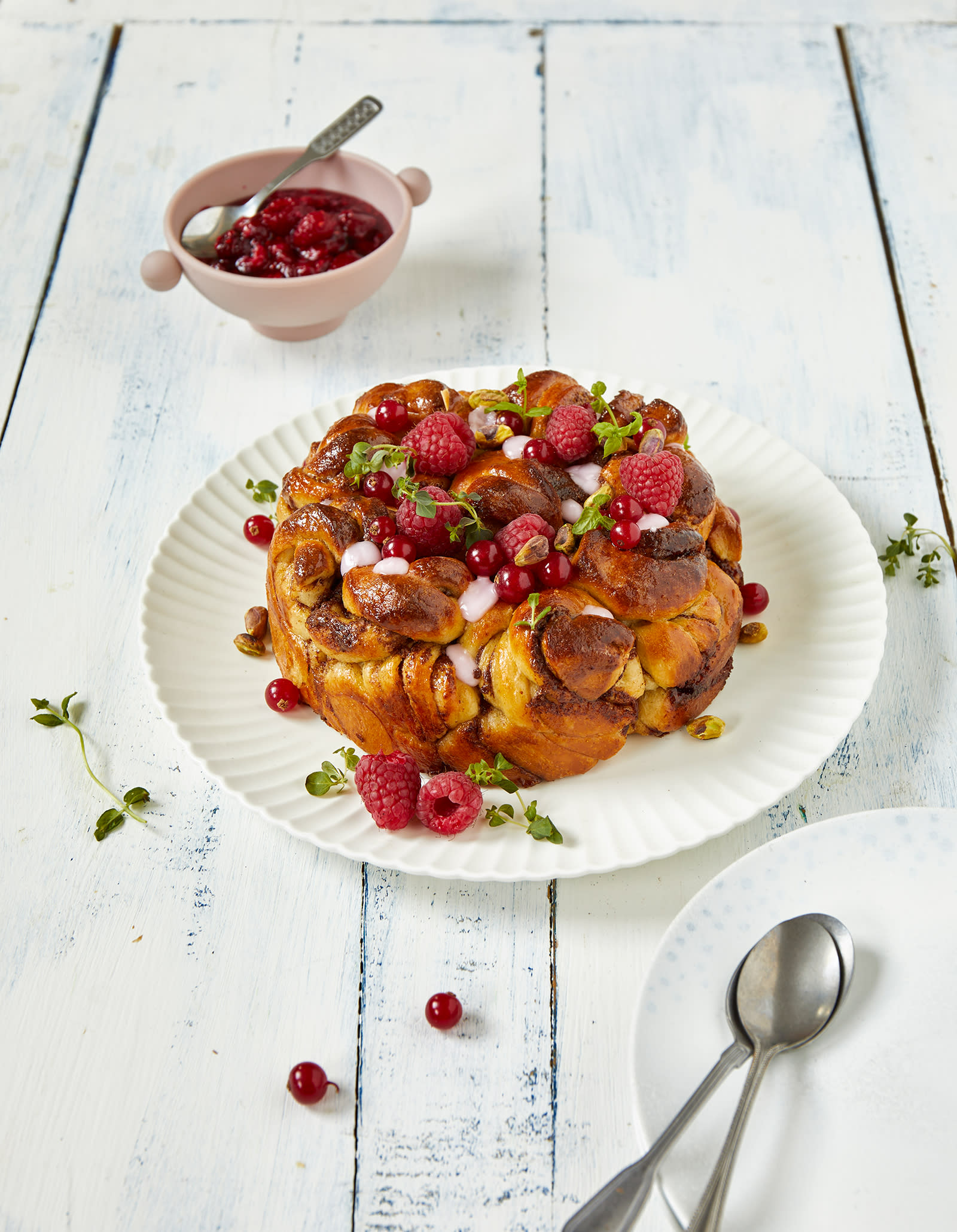 Kanelbollekake med rips og bringebær på toppen, pyntet med friske urter, og servert på hvitt fat på hvitt trebord, med en rosa skål rørte bringebær i bakgrunnen.