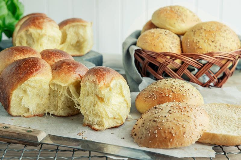 Saftig og luftig brioche i form av loff og rundstykker servert på rist på kjøkkebord av mørkt tre, og i kurv med lyseblått linserviett.