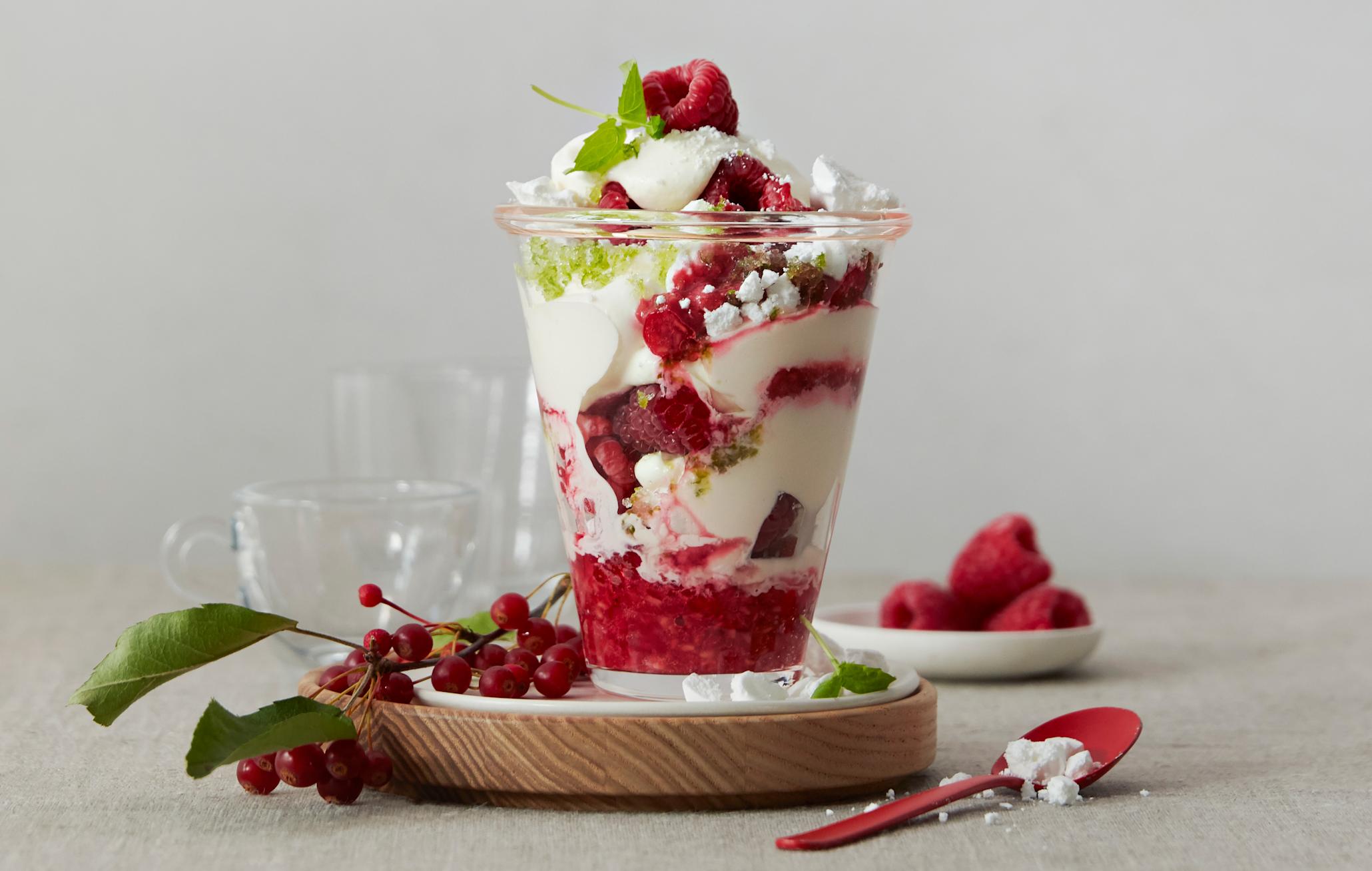 Eton Mess med bringebær, knuste marengs og limesukker servert lagvis i glass på liten treskål. Kvist med grønne blader og røde bær ligger ved siden av, og en rød teskje med knuste marengs ligger i forkant.