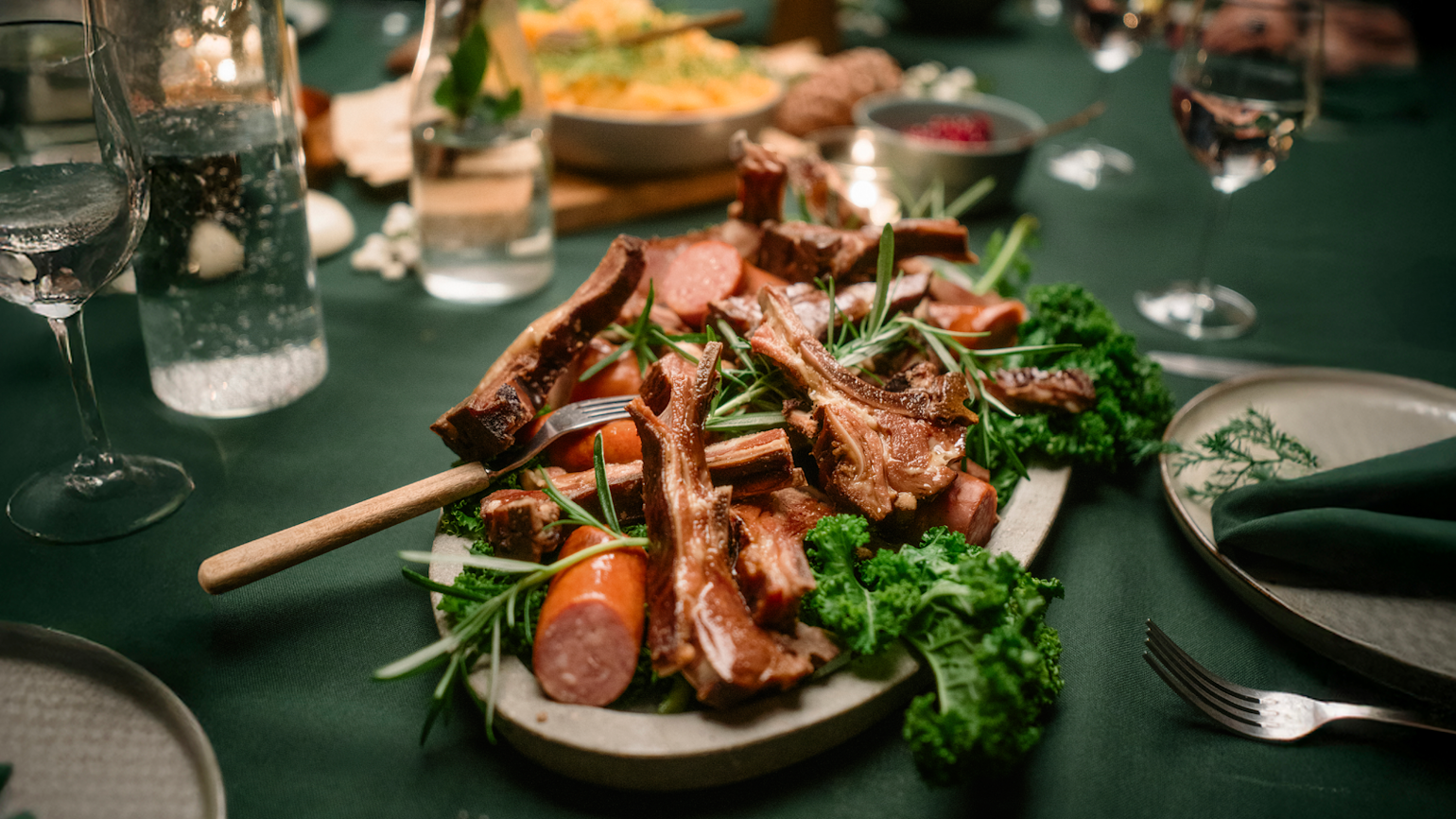 Pinnekjøtt servert på ovalt fat med vossakorv, grønnkål og frisk rosmarin. Fatet står på Mega-grønn duk på dekket julebord.