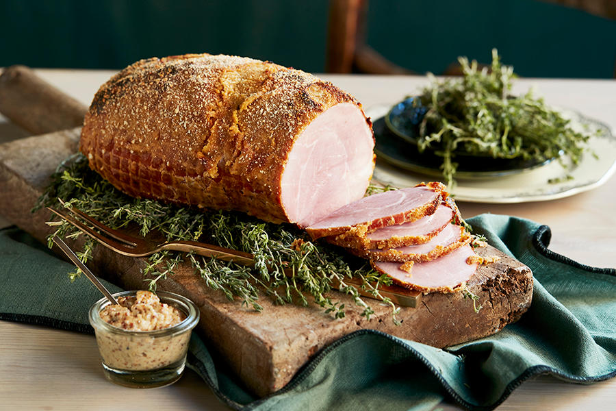 Gratinert juleskinke med sennep, servert på trefjøl på en seng av ferske urter. Liten krukke med grov sennep ved siden av.