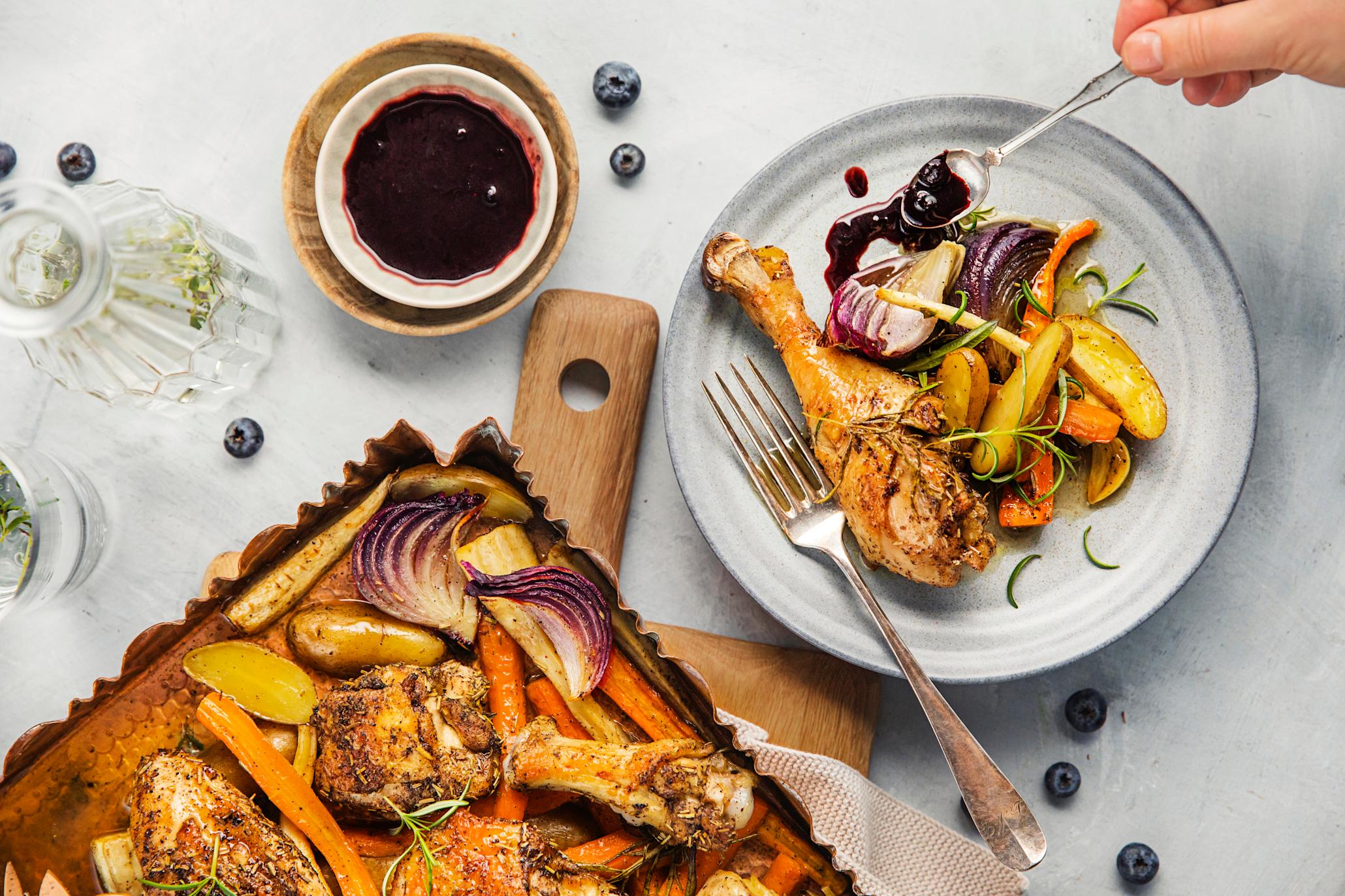 Ovnsbakt kylling med kraftig blåbærsausi langpanne ovenfra ved siden av en tallerken med kylling og rotgrønnsaker på og en hånd som tar bringebærsaus på tallerken