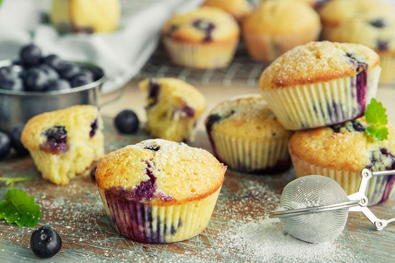 Supersaftige blåbærmuffins drysset med melis og servert på blå rustikk trefjøl. Skål med friske blåbær, og kjølerist med flere muffins i bakgrunnen.