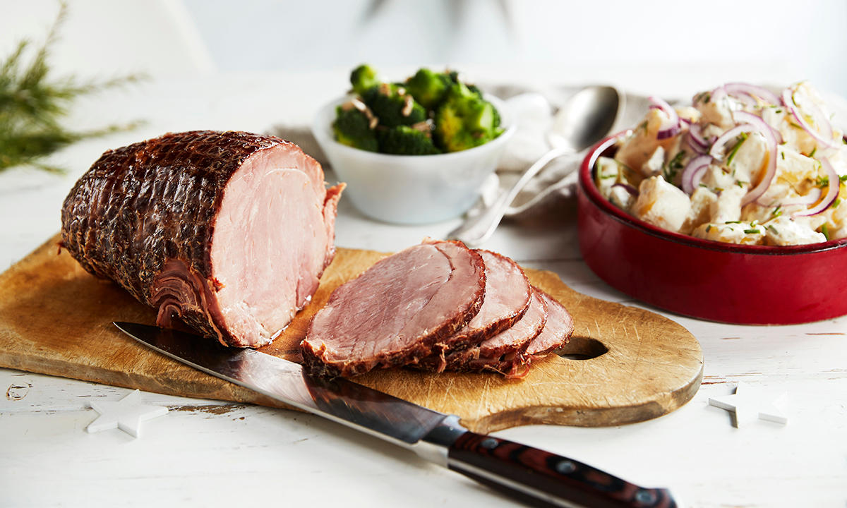 Juleskinke i skiver servert på trefjøl, med rød skål hjemmelaget potetsalat ved siden av. Liten hvit skål med dampet brokkoli i bakgrunnen.