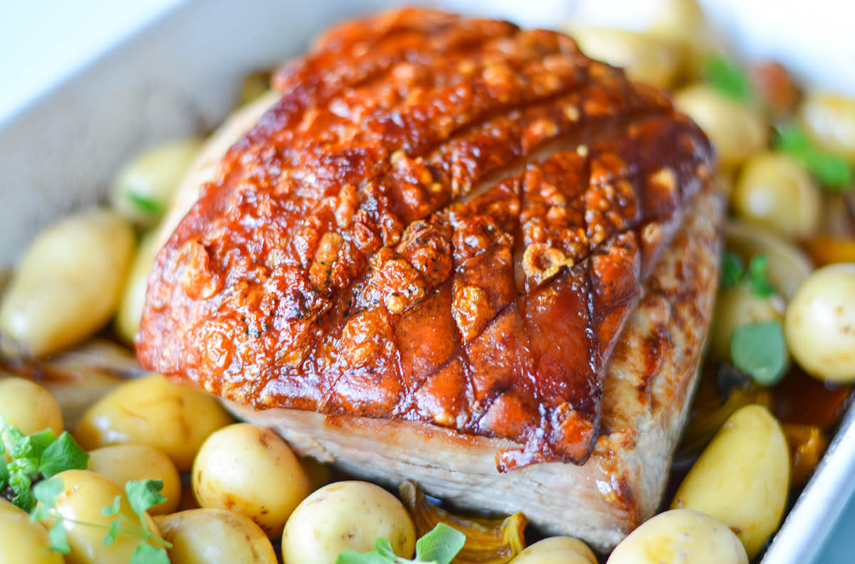 Glasert skinkestek med sprø svor, servert i hvit ildfast form med ovnsbakte poteter, gule beter og løk. Ferske urter er strødd over potetene.