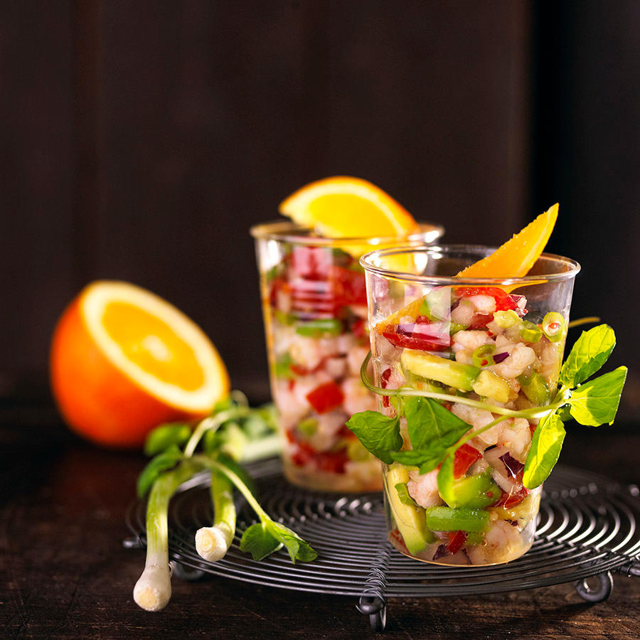 Reker ceviche servert i to gjennomsiktige glass, med avokado, tomater, rødløk, chili og en appelsinskive på toppen. En halv fersk appelsin i bakgrunnen og fersk vårløk på siden.