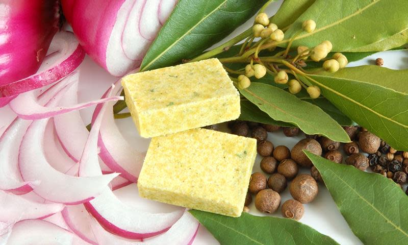 Buljongterninger ligger på et hvitt underlag sammen med skiver av rødløk, laurbærblad, hel pepper og annet krydder.