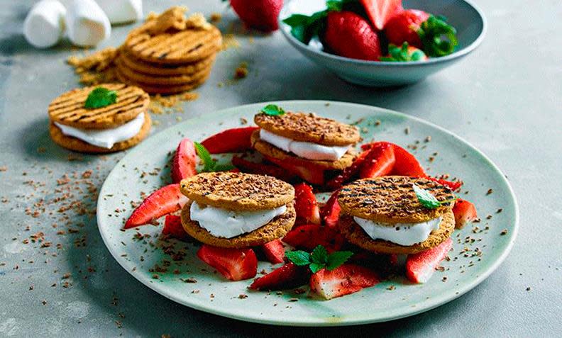 Desserten S'mores; grillet marshmallows med smeltet sjokolade mellom to kjeks, ligger på tallerken med ferske jordbærskiver og jordbærsaus. Ferske jordbær, kjeks, og marshmallows i bakgrunnen.