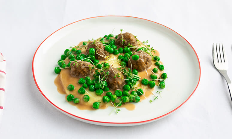 Kjøttboller med potetmos, fløtesaus og erter servert på en hvit tallerken med rød kant som ligger på en hvit duk.