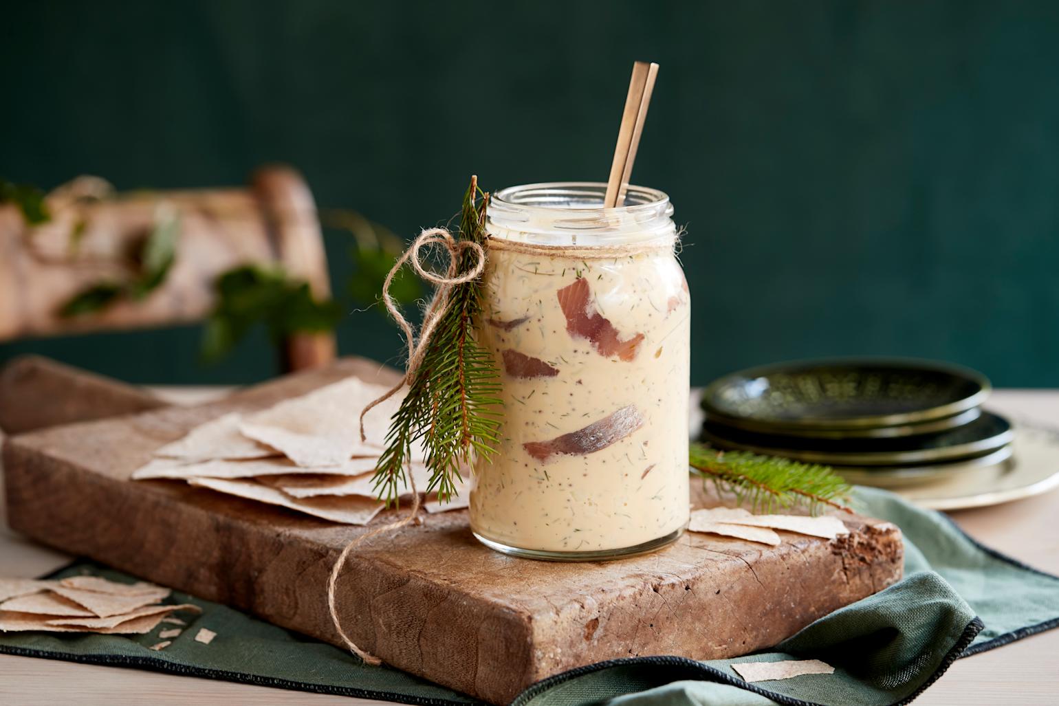 Sennepsild med eple og dill servert i Norgesglass på rustikk trefjøl med flatbrød. Fjøla og glasset er pyntet med granbar, og står på en mørkegrønn stoffserviett.