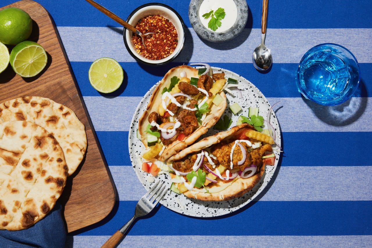 Gyros med syltet hodekål, pommes frites, grønnsaker og gyrossaus servert i pitabrød på prikkete lys tallerken, på blåstripet duk. Små skåler med krydder og saus ved siden av, og skjærefjøl med pitabrød og lime på siden.