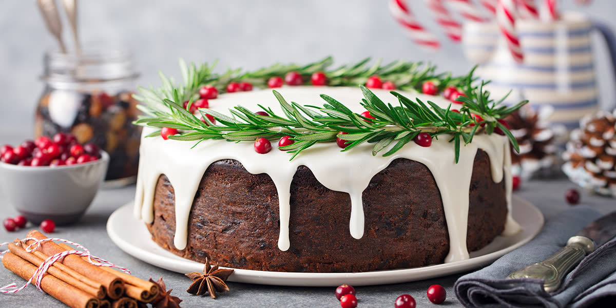 Engelsk fruktkake med royal icing pyntet med rosmarin og tyttebær i form av en julekrans, og servert på en hvit tallerken. Tørket krydder og julepynt rundt.