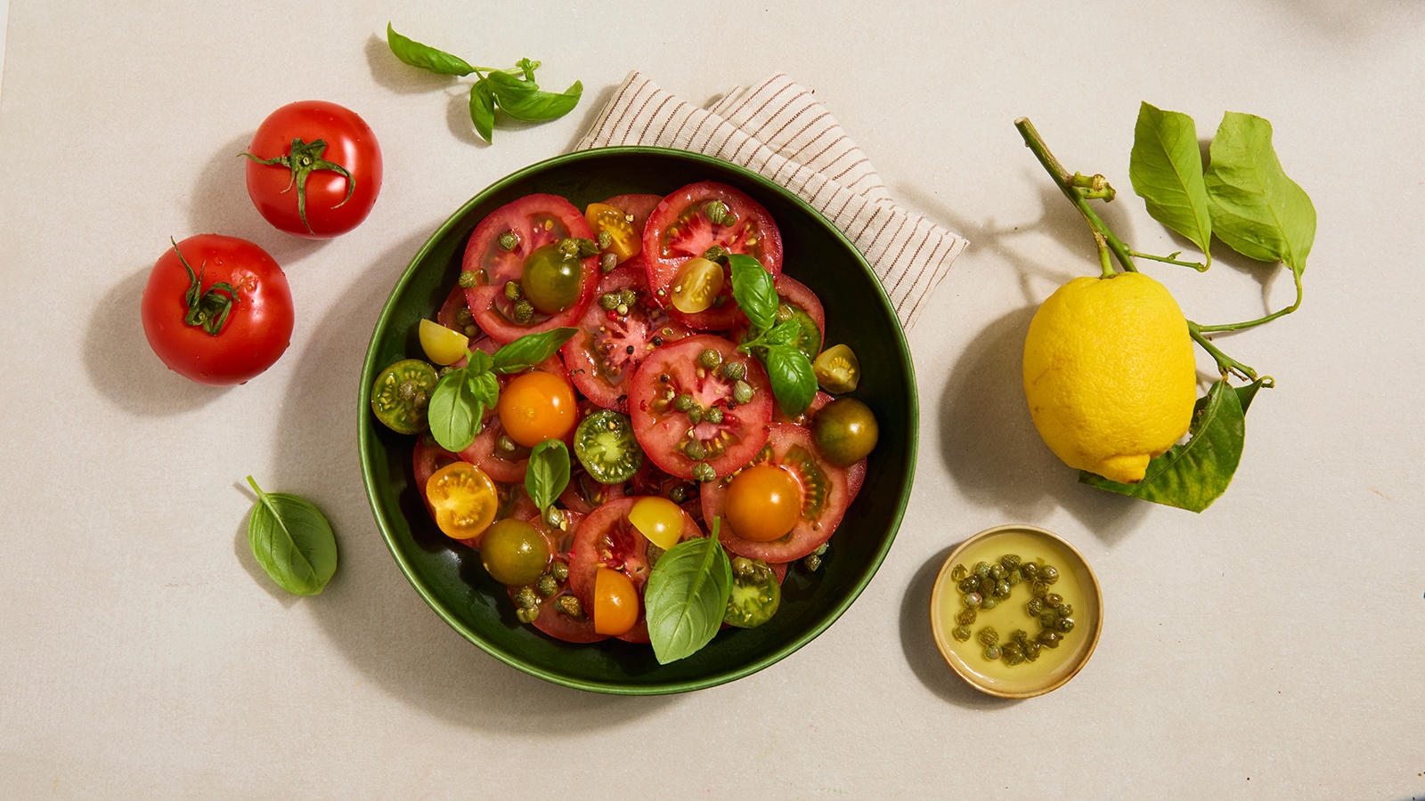 Fargerik tomatsalat med kapers servert i mørkegrønn form med frisk sitron og tomater på siden.