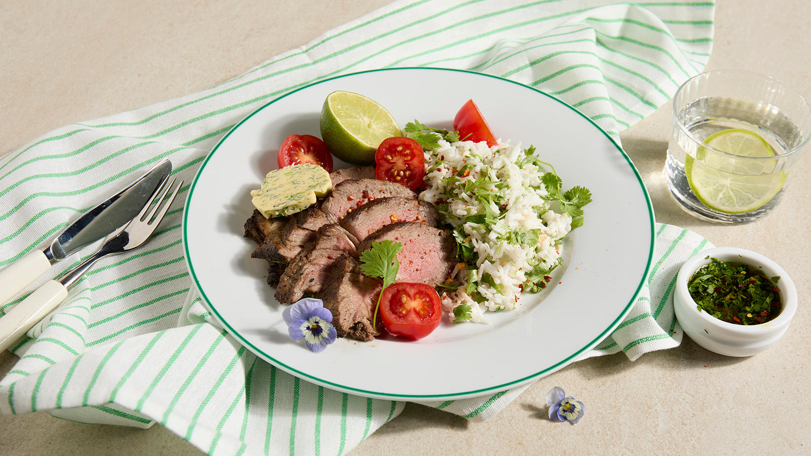 Grillet entrecôte med kokosris og hjemmelaget hvitløkssmør servert med tomater og en halv lime på hvit tallerken med grønn kant.