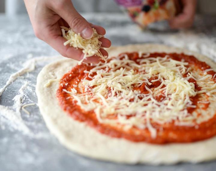 Kjevlet rund pizzadeig på melet underlag, med rød saus, får strødd revet mozzarella over seg.
