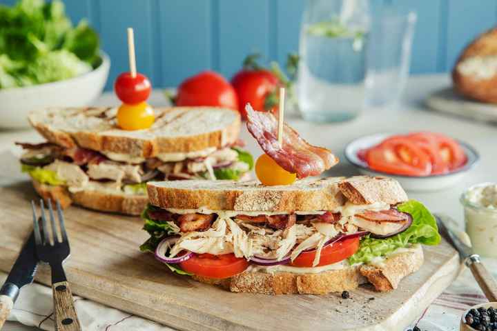 To stykk Club sandwich med grillet kylling, bacon, tomat, salat og rødløk mellom to skiver rustikk landbrød. Servert på trefjøl, med blå bakrunn. Tannpirkere med fargerike tomater stukket ned i hver sandwich.