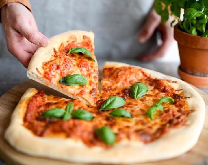 Rund pizza Margherita med rød saus og ost, toppet med fersk basilikum. En hånd i bildet som tar opp et stykke pizza.