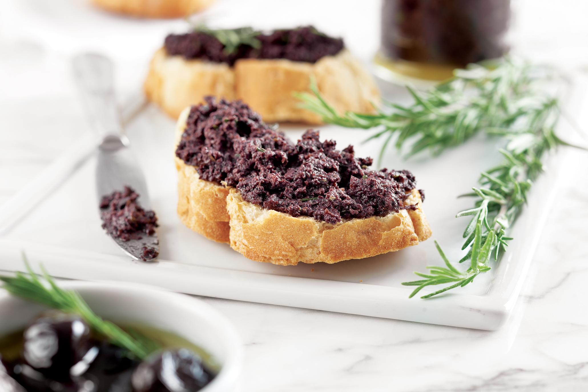 Hjemmelaget oliventapenade på skiver med lyst brød. Servert på hvitt firkantet fat, med ferske urter ved siden av.