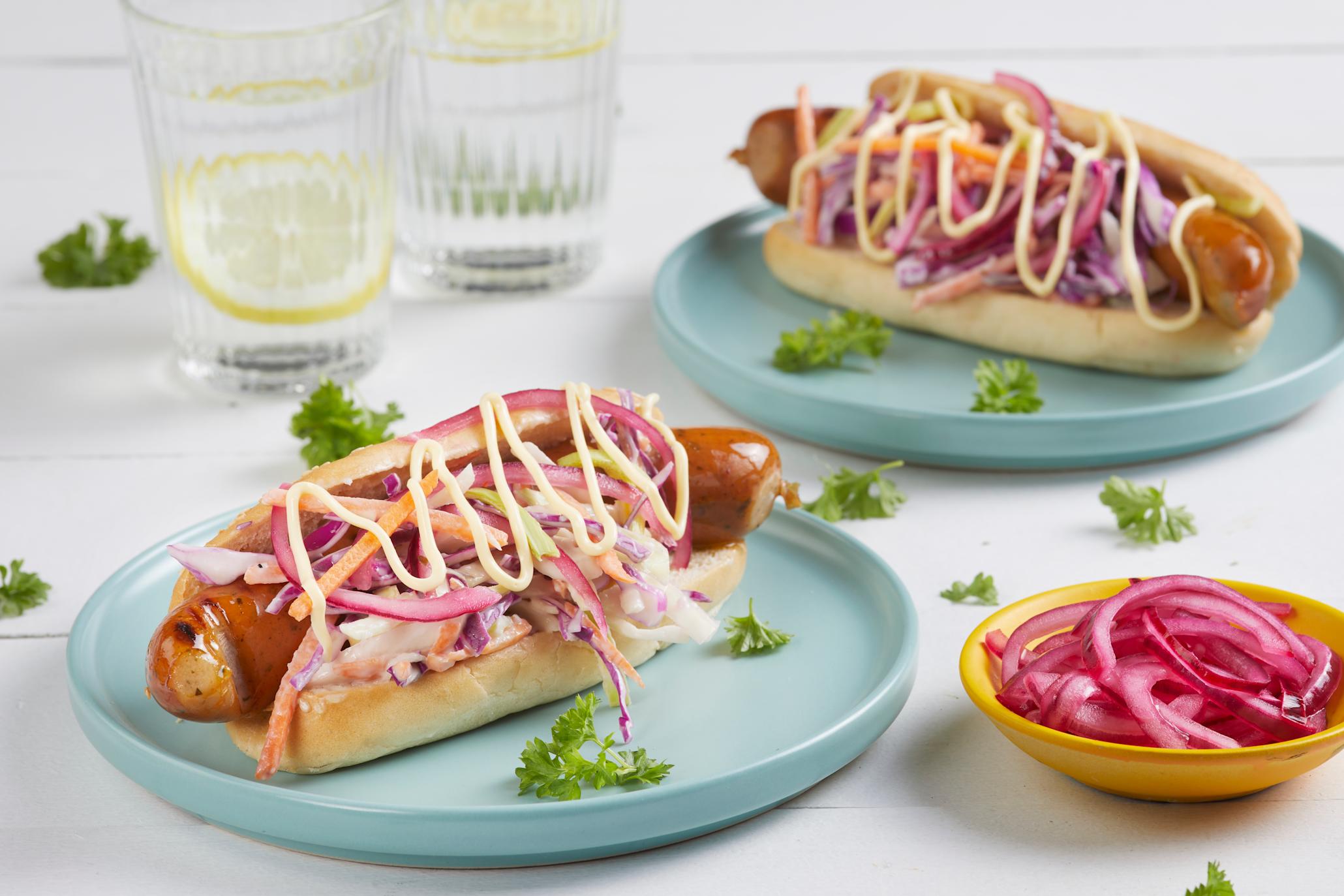 Tasty Garlic pølse med hjemmelaget coleslaw og syltet rødløk servert i pølsebrød på lyseblå asjetter på hvitt bord. Liten gul skål med syltet rødløk ved siden av, og vannglass med sitronskiver i bakgrunnen.