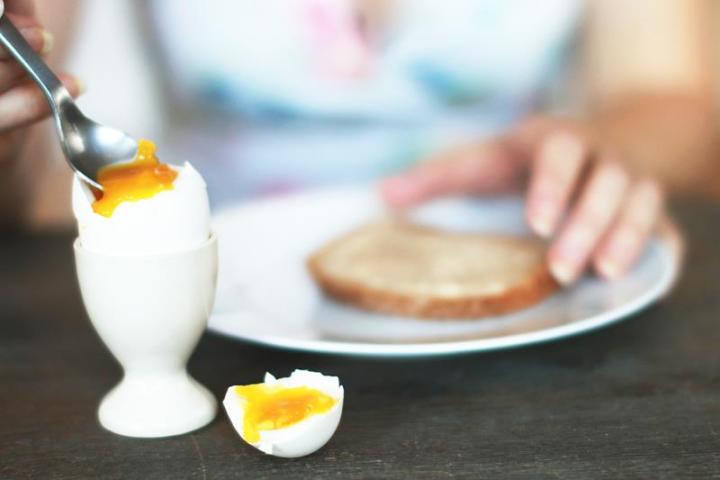 Kokt egg i hvitt eggeglass, på frokostbord med tallerken og brødskive. Toppen er skåret av det kokte egget.